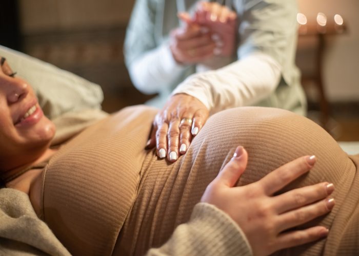 Cropped image of smiling pregnant woman and midwife at home. Woman in casual clothes lying on bed, midwife holding hand. Pregnancy, medicine, home birth concept